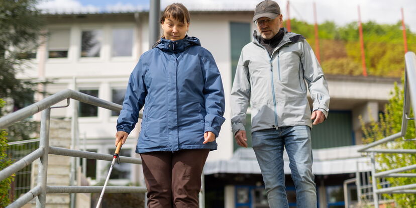 Eine Frau mit einem Langstock steht an einem Treppengeländer, ein Lehrer für O&M steht neben ihr.