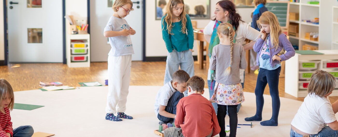 Einblick in die Lernräume. Eine Lernbegleiterin umringt von einer Gruppe Kinder.