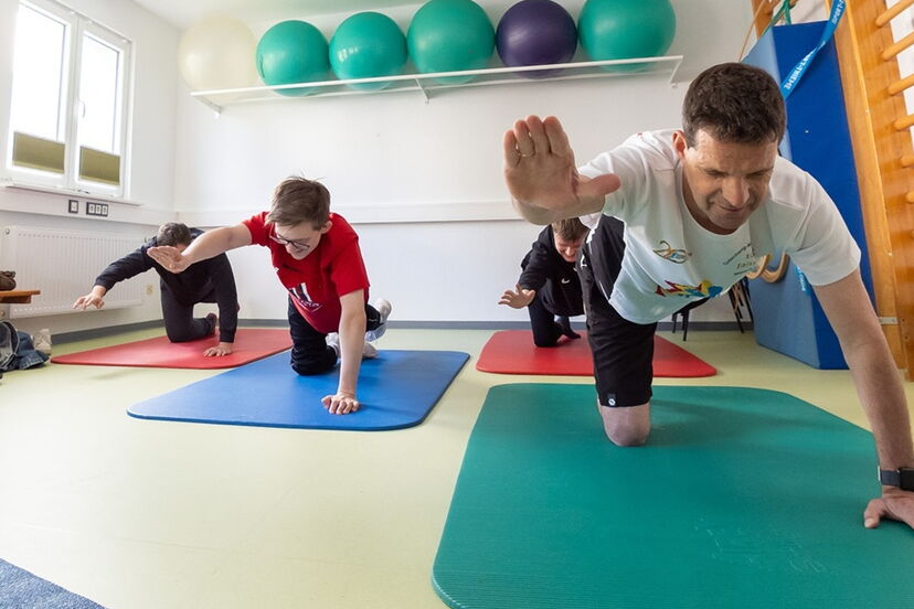 Vier Teilnehmer und eine Motologin machen eine Gymnastikübung, bei der sie jeweils ein Arm und ein Bein im Vierfüßlerstand ausstrecken.