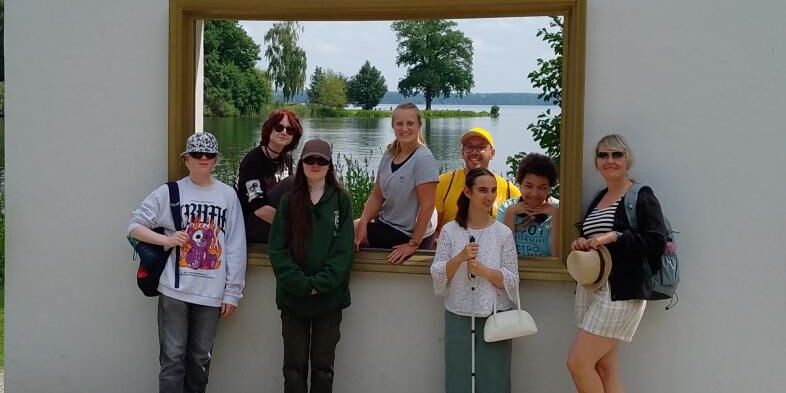 6 Schüler*innen und zwei Betreuer*innen stehen vor einem Fenster, das auf ein Ufer mit Bäumen und Wasser zeigt und scheinen sichtlich ihren wohlverdienten Urlaub zu genießen.