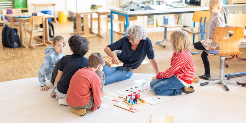 Eine Lernbegleitung sitzt mit Schüler*innen auf dem Boden und demonstriert Material.