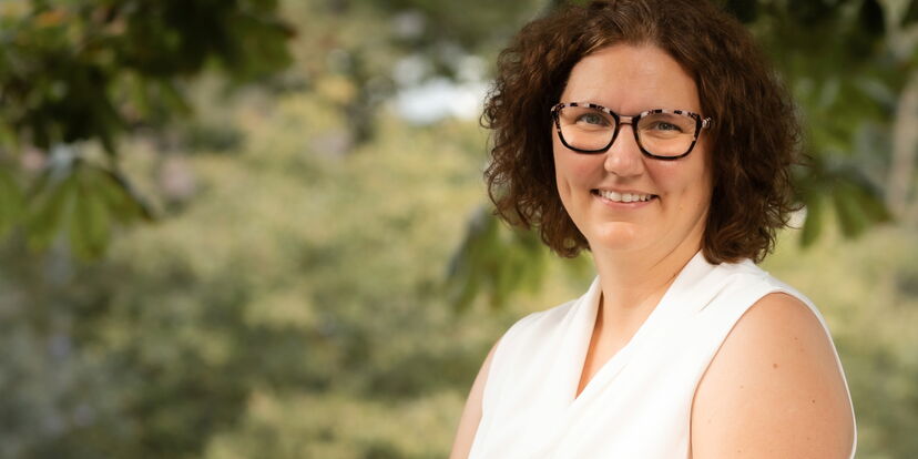 Die Schulleiterin der Montessori Schule lächelt in die Kamera. Sie hat braune schulterlange kleine Locken und trägt eine Brille. Sie trägt ein weißes Oberteil.
