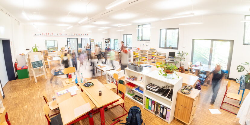 Ein Lernraum mit Möbeln, Ranzen und vielen sich bewegenden Menschen.
