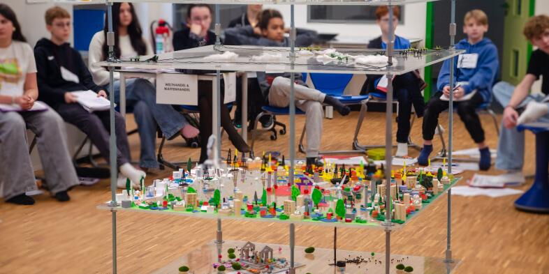 Das vierstöckige, große Spielboard mit diversen Figuren steht in der Mitte der Aula, ringsum sitzen Schüler*innen auf Stühlen.