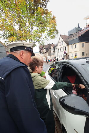 Gemeinsam mit einem Polizisten überreicht ein blista-Schüler einer Person in einem Auto eine grüne Karte für rücksichtsvolles Fahrverhalten.