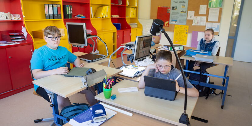 Drei Schüler*innen sitzen an ihren Arbeitsplätzen und arbeiten mit unterschiedlichen Medien.