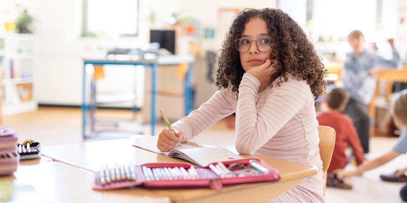 Im Unterrichtsgeschehen sitzt ein Mädchen an einem Tisch. Sie hat einen Stift in der Hand und scheint nachzudenken.