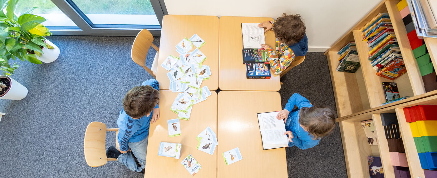 In einem Arbeitsraum blickt man von oben auf einen Tisch, an dem drei Kinder sitzen und mit unterschiedlichen Materialien arbeiten.