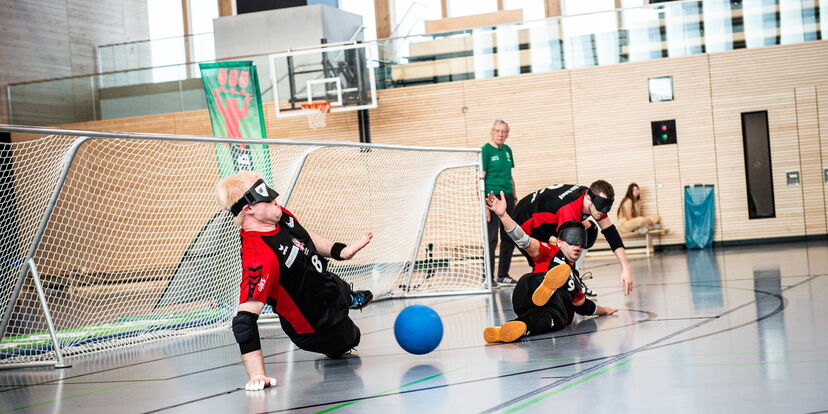 In einer Sporthalle werfen sich drei Goalballspieler vor ein Tor, um einen Ball abzuwehren.