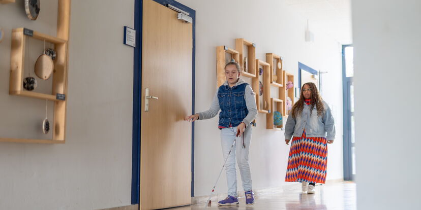 Eine Schülerin mit Langstock läuft unter den Augen einer Reha-Lehrerin durch ein Gebäude.