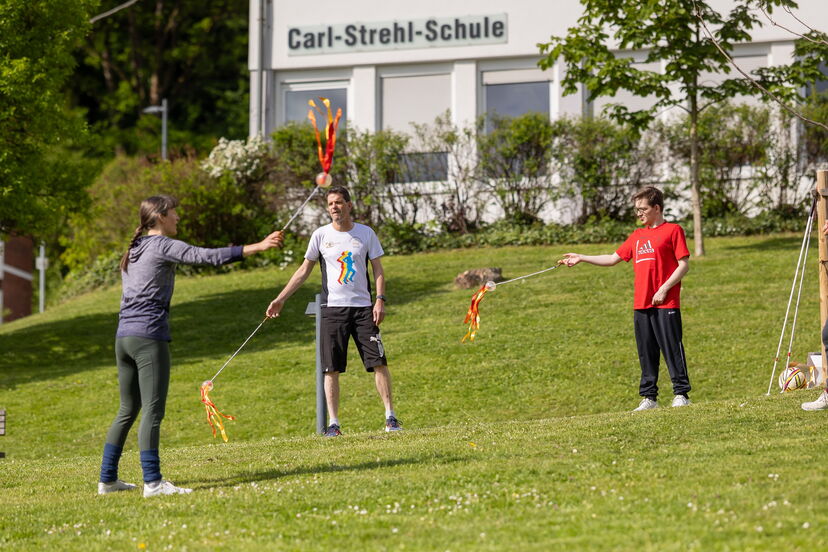 Personen in Sportkleidung spielen auf einer Wiese auf dem blistaCampus und schwingen lange Stäbe mit bunten Bändern an den Enden.