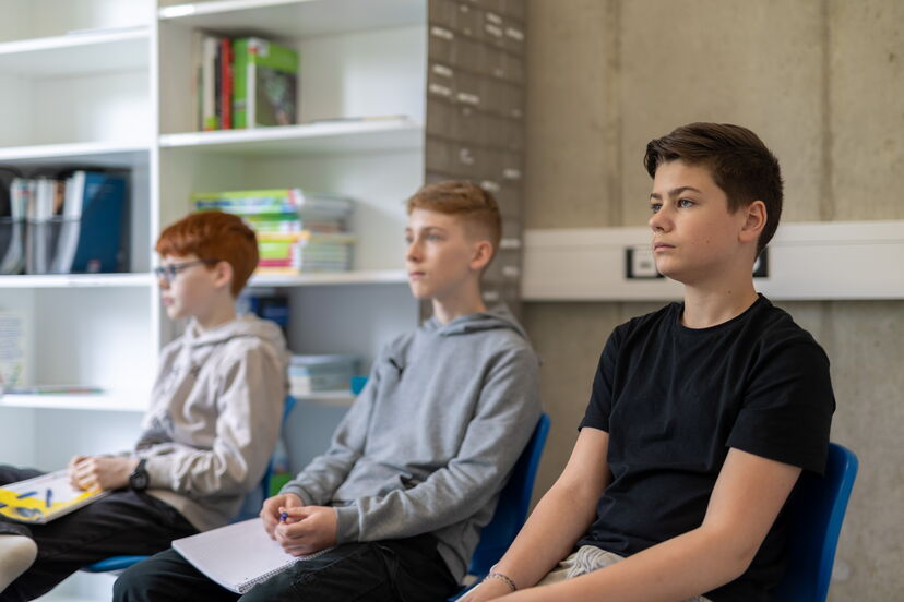 Drei Schüler sitzen auf Stühlen und haben Schreibblöcke auf dem Schoß.