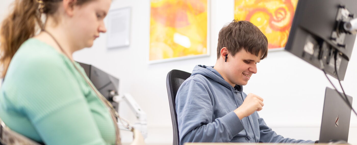 Zwei Azubis sitzen in einem Raum mit Laptops und Bildschirmen.