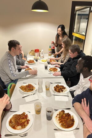 Die Gruppe sitzt an einem langen Tisch, vor sich Teller mit Nudeln und Becher mit Getränken.