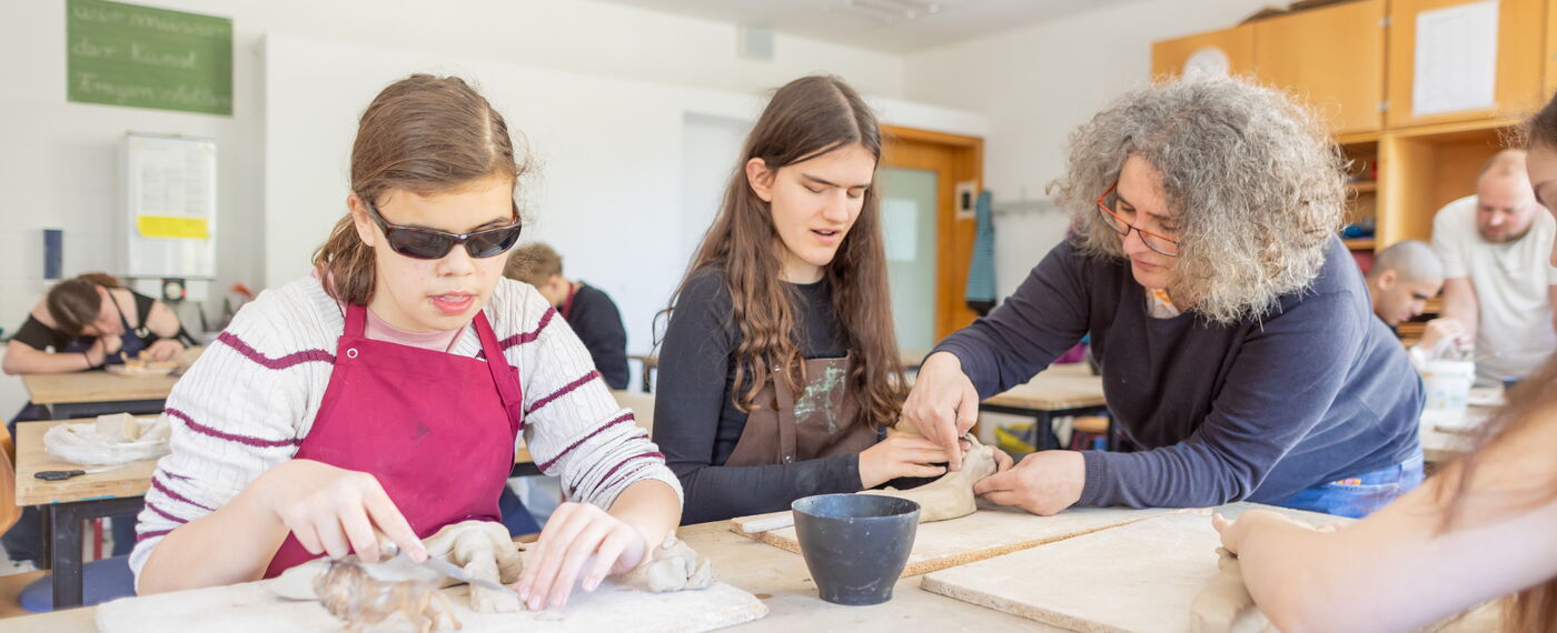 Zwei Schülerinnen formen Tiere aus ton, während eine Lehrerin etwas daran ertastet.