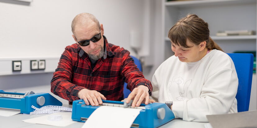 Zwei Menschen sitzen vor Punkschriftmaschinen, ein Mann lehnt sich zu der Punktschriftmaschine einer Frau hinüber.