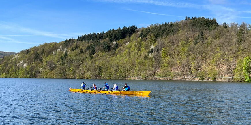 Sechs Personen sitzen in einem gelben Ruderboot auf einem See, im Hintergrund ein frühlingshafter Wald.
