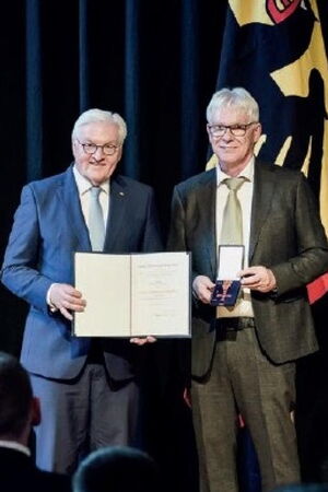 Steinmeier und Duncker stehen nebeneinander und halten die Urkunde und den Orden lächelnd in die Kamera.