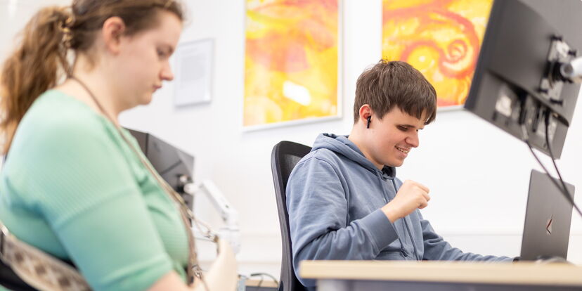 Zwei Azubis sitzen in einem Raum mit Laptops und Bildschirmen.