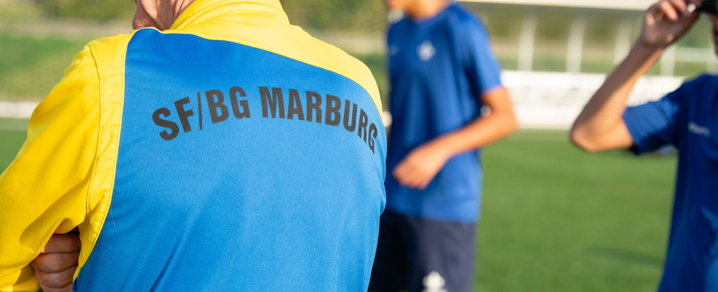 Während des Fußballtrainings sieht man den Rücken des Trainers mit Trainingsjacke im Vordergrund, während die Spieler im Hintergrund trainieren.