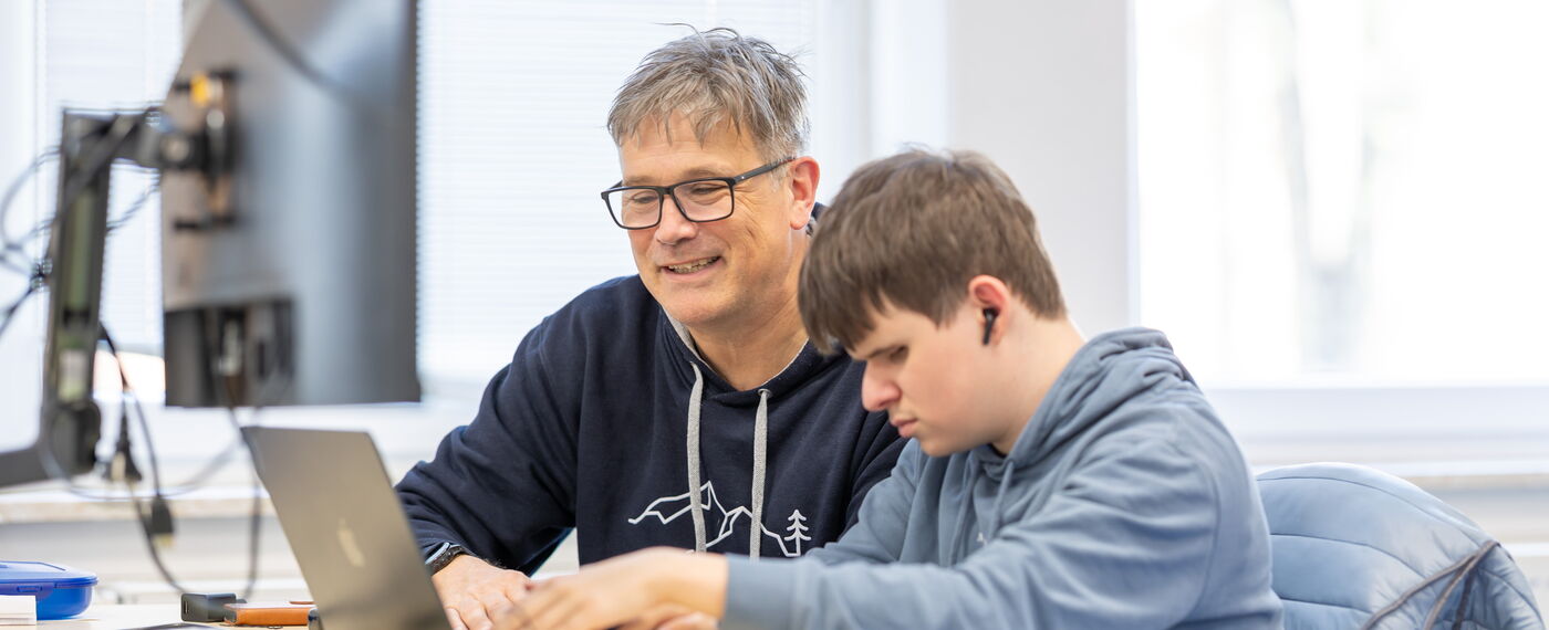 Eine Lehrkraft und ein Auszubildener sitzen an einem Laptop, der Azubi arbeitet mit einen Screenreader .