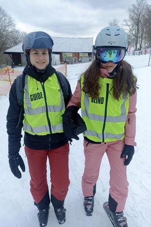 Guide führt Schülerin im Schnee, beide Frauen tragen Skikleidung und Skischuhe, im Hintergrund ist eine Hütte zu sehen.