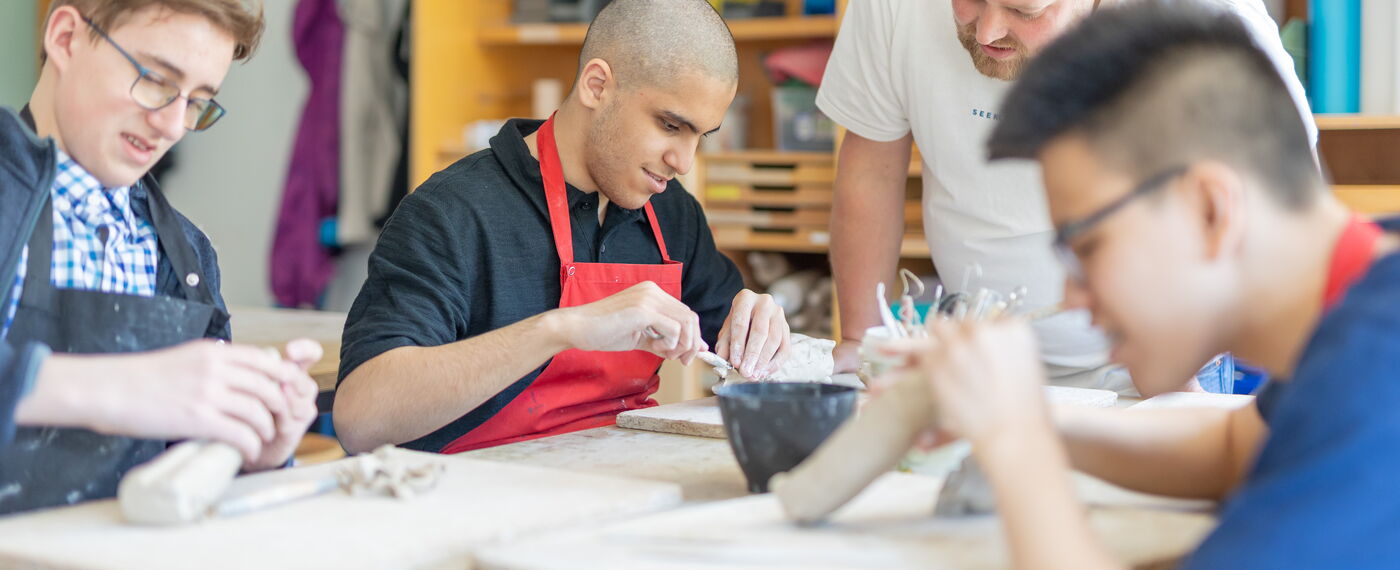 Eine Klasse formt etwas aus Ton, eine Lehrkraft schaut interessiert.