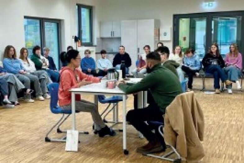 In der Aula der Montessori-Schule sitzen drei Menschen an einem Tisch, die Schüler*innen sitzen ringsum im Stuhlkreis