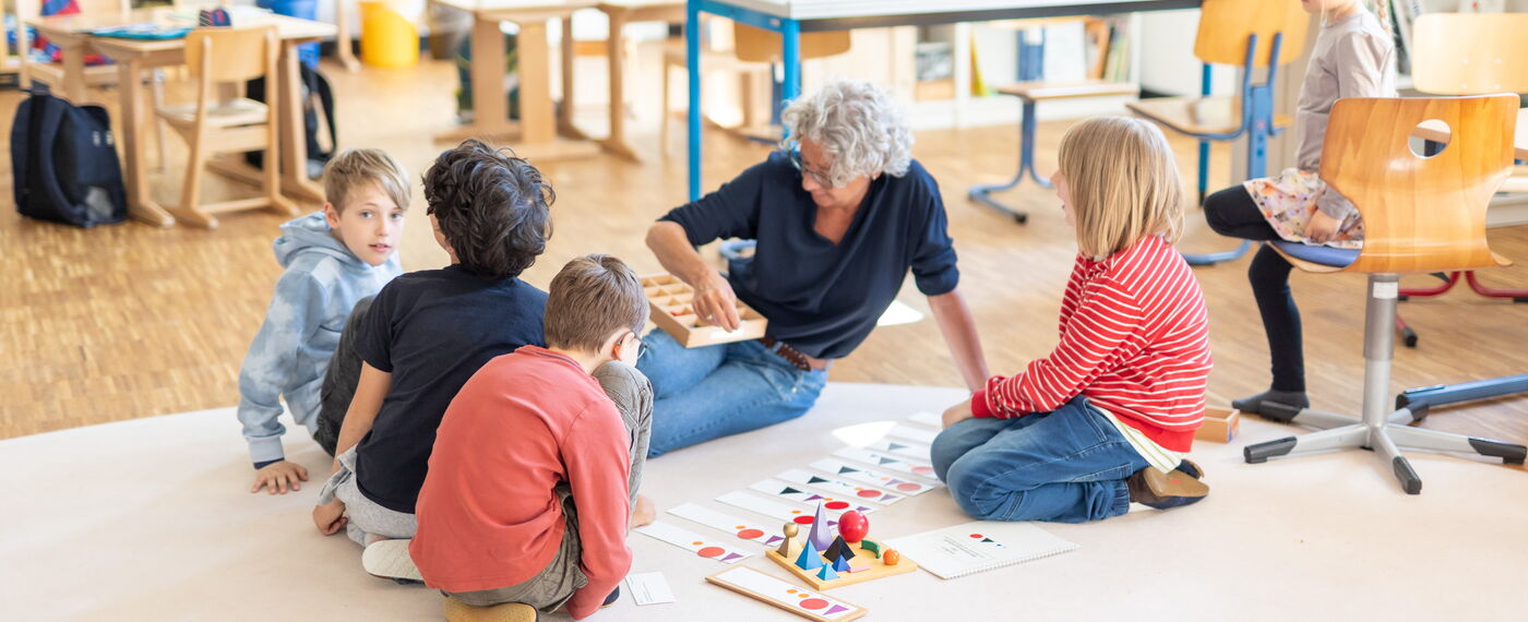 Eine Lernbegleitung sitzt mit Schüler*innen auf dem Boden und demonstriert Material.