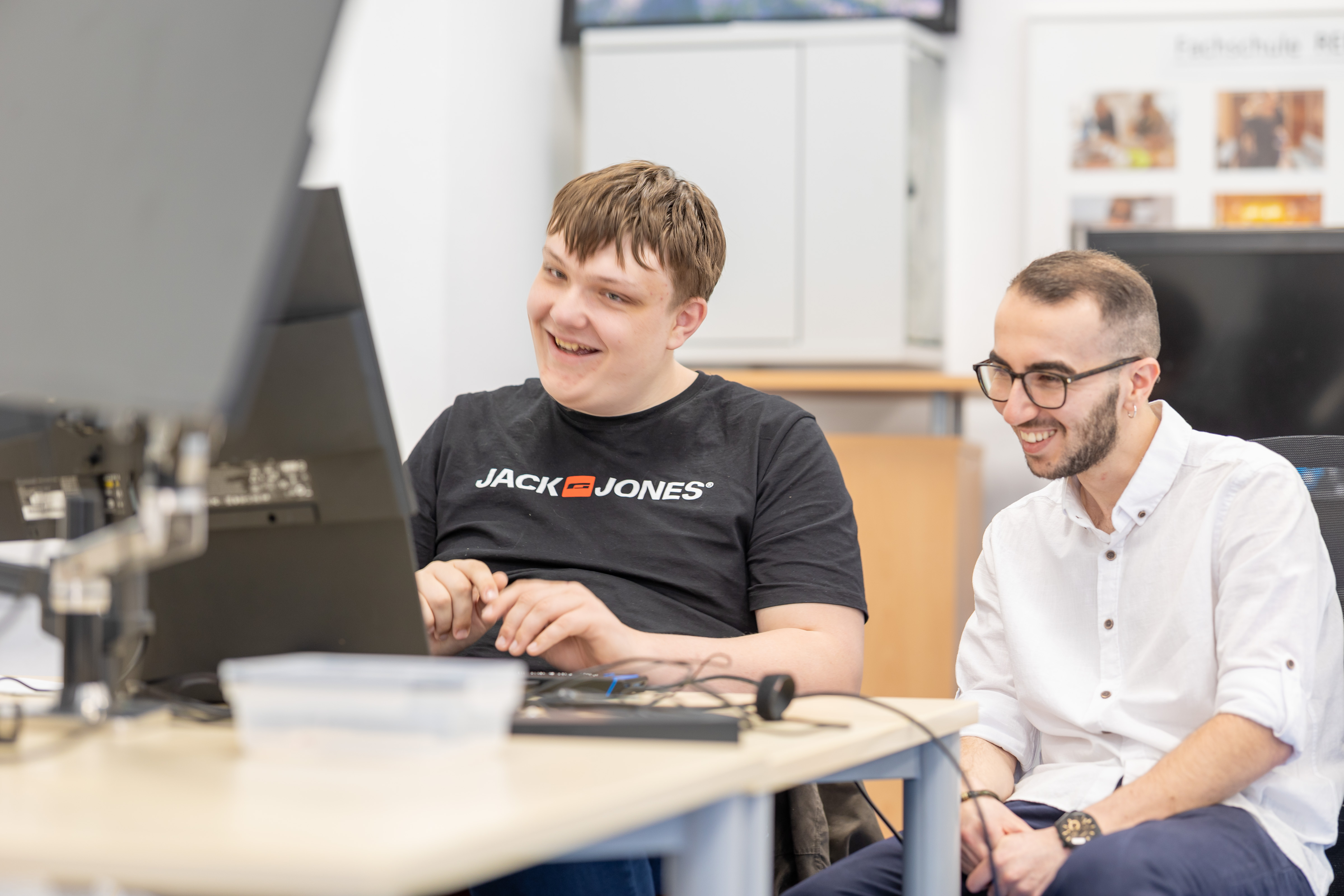 Zwei junge Männer sitzen gemeinsam an einem Pc und lächeln.